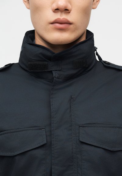 Gros plan sur une personne portant une veste sombre à col montant avec fermeture Velcro et deux poches à rabat sur la poitrine.