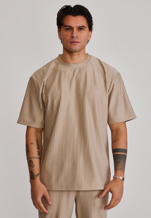 Mies, jolla on beigeä ribbineulosta oleva lyhythihainen paita ja siihen sopivat housut, näyttää tatuoidut käsivarret ja hopeisen rannekorun oikeassa ranteessa.