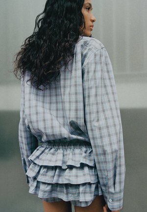 Femme aux longs cheveux bouclés portant une jupe à volants à carreaux bleu clair et une chemise oversize à manches longues assortie, de profil.