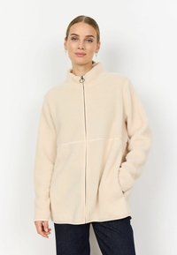 Beige fleece-vetoketjullinen takki, jossa on seisomapanta, sujuva vetskari ja sivutaskut, suunniteltu pehmeästä, teksturoidusta kankaasta.