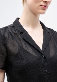 Κοντινό πλάνο ενός ατόμου που φοράει διάφανη μαύρη κοντομάνικη μπλούζα με γιακά και κουμπιά, εμφανίζοντας το ντεκολτέ και το άνω μέρος του κορ μού.