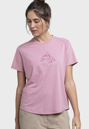 Frau mit zurückgebundenen Haaren, die ein hellviolettes T-Shirt mit einem pinken und weißen abstrakten Design trägt, steht vor einem einfarbigen Hintergrund.