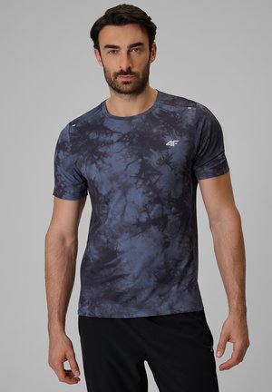 Mężczyzna w ciemnoniebieskiej i czarnej koszulce tie-dye z krótkim rękawem i małym białym logo na piersi, stojący na szarym tle.