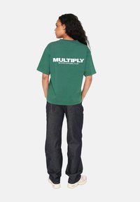 Camiseta de algodón verde con escote redondo, que presenta el texto blanco "MULTIPLY APPAREL CLUB" en la parte de atrás. Combinada con jeans de mezclilla oscura.