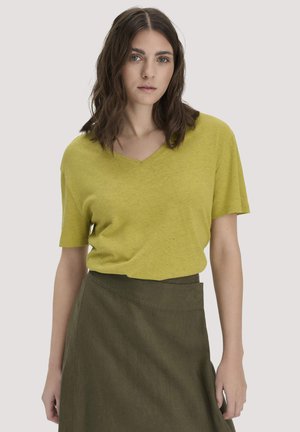 Vrouw met bruin haar tot op de schouders, gekleed in een lichtgroen V-hals shirt met korte mouwen en een olijfgroene rok, tegen een effen achtergrond.