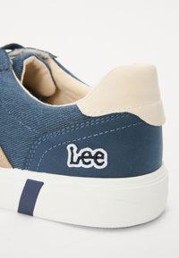 Baskets en toile bleue avec des accents en cuir beige, dotées d'une semelle en caoutchouc blanche et du logo "Lee" brodé en blanc.