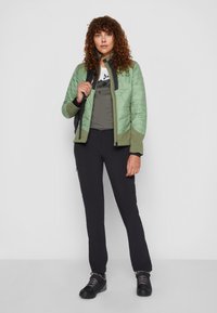 Green quilted jacket with a high collar, paired with gray t-shirt and black pants. Model wears black hiking boots and carries a backpack.