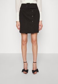 Žena v černé mini sukni s páskem a knoflíky v kombinaci s černými sandály na vysokém podpatku zdobenými velkými mašlemi.