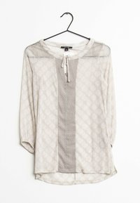 Blouse beige clair à manches longues avec un motif discret de feuilles et un panneau vertical texturé central, suspendue à un cintre noir sur fond blanc.