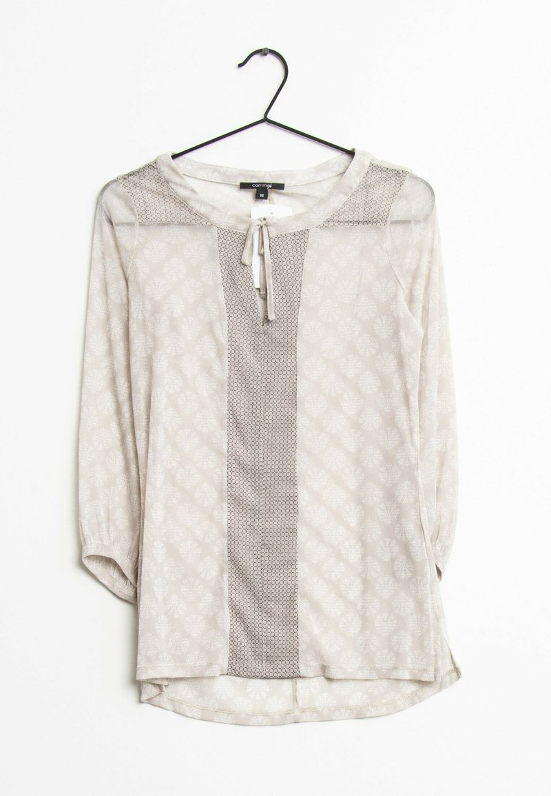 Blouse beige clair à manches longues avec un motif discret de feuilles et un panneau vertical texturé central, suspendue à un cintre noir sur fond blanc.