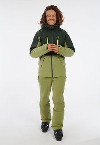 Zelená a tmavozelená izolovaná bunda so zipsovými vreckami, skombinovaná so zelenými nohavicami. Model stojí v lyžiarskych topánkach na bielom pozadí.