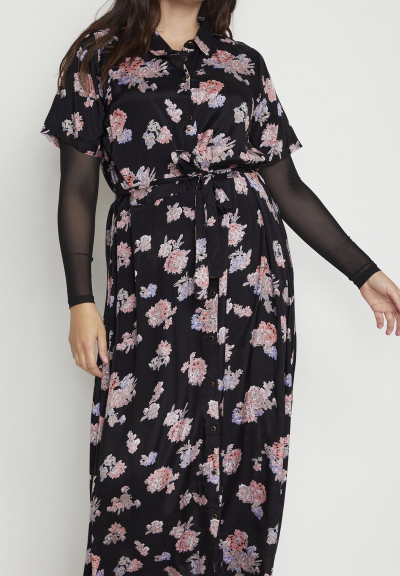 Femme portant une robe noire à motifs floraux avec des boutons sur le devant, une ceinture à la taille et un top noir transparent à manches longues en dessous, debout sur un fond blanc.