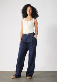 Bijela rebrasta majica bez rukava s detaljem gumba, uparena s visokim tamnoplavim traper hlačama širokih nogavica. Neutralni baletanke upotpunjuju look.