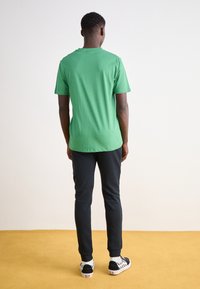 Man som bär en grön t-shirt, svarta byxor och rutiga sneakers står med ryggen mot kameran på ett gult golv och en vit vägg i bakgrunden.