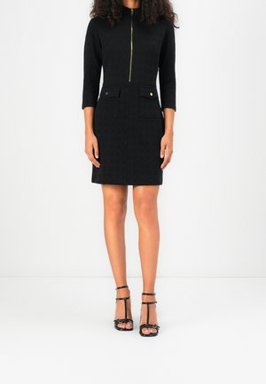 Žena v černých šatech po kolena s tříčtvrtečními rukávy, předním zipem, dvěma předními kapsami a černými sandály na podpatku se zapínáním přes nárt.