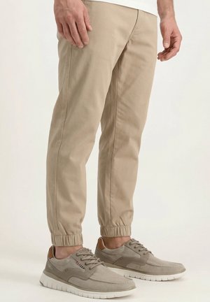 Mężczyzna w beżowych spodniach dresowych i taupe sznurowanych trampkach, stojący na białej powierzchni na jednolitym tle.