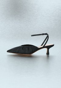 Μαύρη μυτερή γόβα με τακούνι kitten, με ραβδωτό ύφασμα και λεπτό ρυθμιζόμενο λουράκι στον αστράγαλο, σε ανοιχτό γκρι υπόβαθρο.