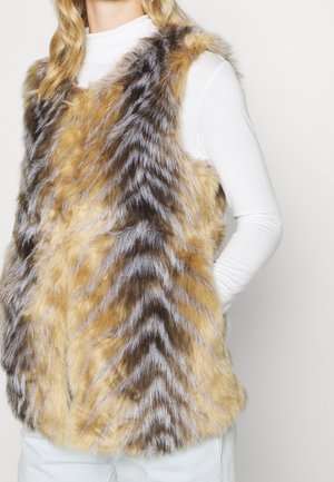 Femme portant un gilet sans manches en fausse fourrure à motif chevron dans des tons marron, noir et gris sur une chemise blanche à manches longues.