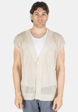 Giovane uomo che indossa un cardigan beige senza maniche a maglia aperta sopra una camicia bianca, in piedi davanti a uno sfondo chiaro e uniforme.