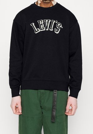 Osoba nosząca czarną bluzę Levi's z białym logo i zielone spodnie z czarnym paskiem Levi's zwisającym z pasa.