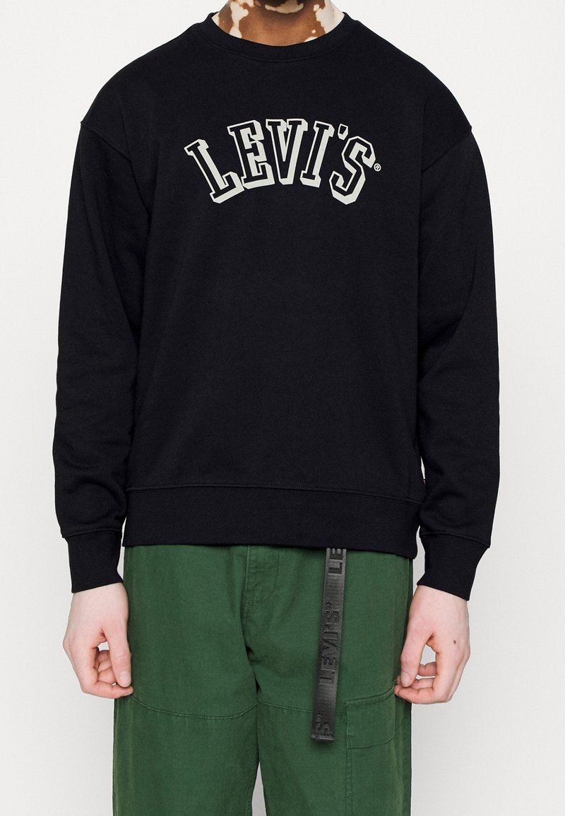 Personne portant un sweat Levi's noir avec un logo blanc et un pantalon vert avec une lanière noire Levi's pendue à la taille.