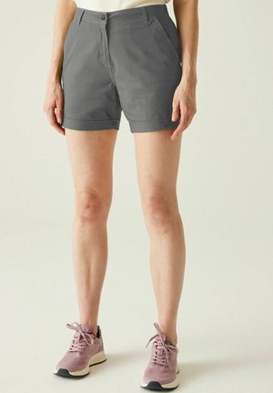 Persona con pantalones cortos grises y zapatillas rosas de pie frente a un fondo claro y liso, mostrada desde la cintura hasta los pies.