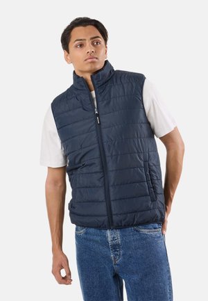 Ung mand iført en navy blå polstret vest over en hvid t-shirt og blå jeans, stående mod en ensartet lys baggrund.