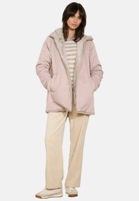Giacca puffer rosa chiaro con cappuccio in pelliccia grigia morbida, tasche frontali e chiusura con zip, indossata sopra un maglione a righe e pantaloni in velluto a coste.