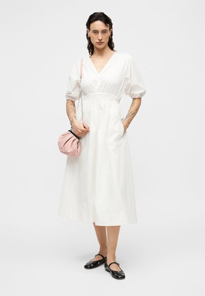 Λευκό βαμβακερό φόρεμα με κοντά φουσκωτά μανίκια, σφηνοειδή μέση και πλαϊνές τσέπες. Το μοντέλο κρατάει ροζ τσάντα και φοράει μαύρα φλατ παπούτσια.