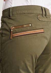 Gros plan sur un pantalon olive avec une poche arrière zippée mettant en avant des détails de rayures tissées marron, orange, blanc et violet.