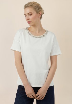 Frau mit blondem Haar, die ein weißes kurzärmeliges Oberteil mit dekorativem Perlenbesatz am Ausschnitt trägt und vor einem hellbeigen Hintergrund posiert.