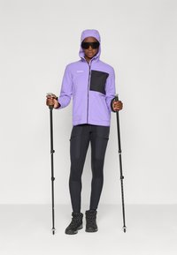 Purple fleece jacket with a black chest pocket, paired with black leggings and hiking boots. Participant holds trekking poles.