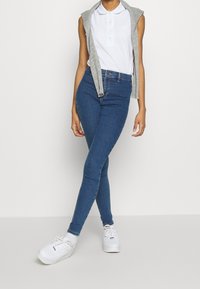 Jeans ajustados de mezclilla azul de talle alto, combinados con una camisa polo blanca y un chaleco sin mangas gris; unas zapatillas blancas completan el look.
