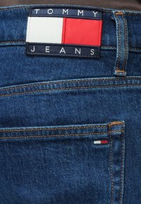 Džinsa bikses ar zilu nokrāsu un oranžu šuvi. Tajās ir melns un sarkans auduma etiķete ar uzrakstu "TOMMY JEANS" un tricolor akcents.