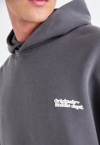 Hoodie en coton gris avec une poche kangourou à l'avant et un capuchon avec cordon de serrage. "Originals Studio dept." est brodé en blanc sur la poitrine.