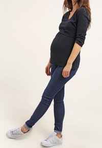 Černý dlouhý rukáv těhotenský top, design s výstřihem do V, nošený s tmavě modrými skinny džínami a bílými teniskami. Hladká tkanina, přiléhavý střih.