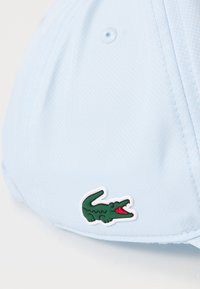 Boné de beisebol azul claro feito de tecido texturizado, apresentando um logo de crocodilo verde com borda branca na frente.
