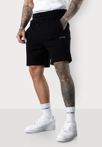 Shorts noirs avec taille à cordon, poches latérales et détails de coutures visibles. Associés à des baskets blanches et des chaussettes mi-hautes.