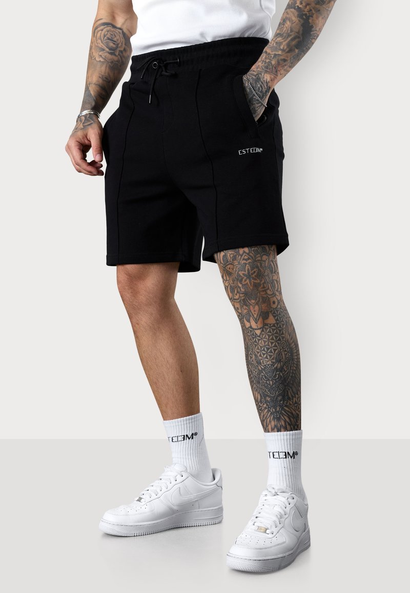 Shorts noirs avec taille à cordon, poches latérales et détails de coutures visibles. Associés à des baskets blanches et des chaussettes mi-hautes.