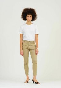 Witte t-shirt met korte mouwen, gecombineerd met beige slim-fit broek en zwarte sandalen met banden. Het model heeft een afro kapsel en staat op een witte achtergrond.