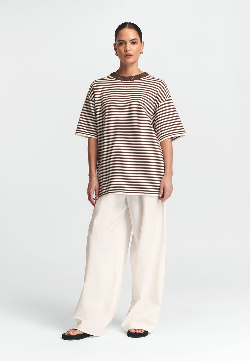 Gestreiftes braunes und cremefarbenes Oversized-T-Shirt mit kurzen Ärmeln, kombiniert mit weißen weiten Hosen und schwarzen flachen Sandalen.