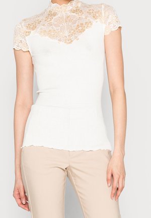 Hvid top med korte ærmer med beige blomsterblonde ved halsudskæring og ærmer, båret med beige bukser på en stående person.