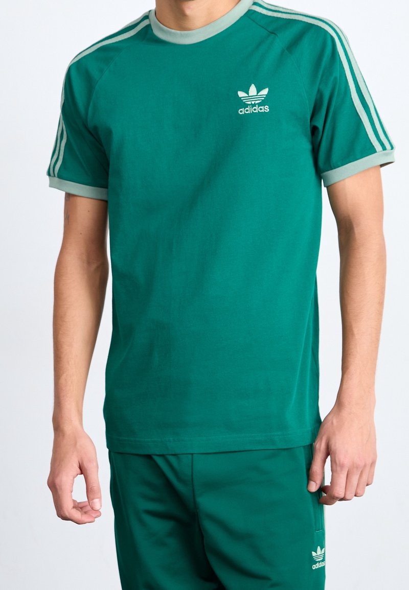 Grön bomullströja med rund hals, korta ärmar och ljusgröna detaljer. Har Adidas-logotypen och tre ränder längs axlarna.