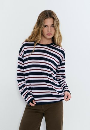 Mujer joven con cabello largo y rubio, vestida con una camiseta de manga larga a rayas azul marino, blanca y granate, y pantalones marrones, frente a un fondo liso y claro.