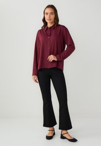 Femme avec des cheveux bruns mi-longs portant un chemisier à manches longues bordeaux à boutons, un pantalon noir évasé et des ballerines noires avec une bride.