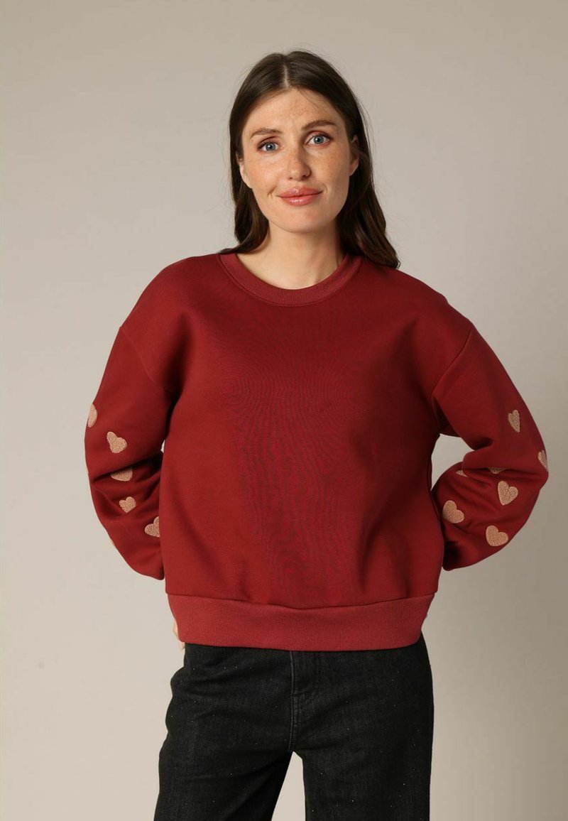 Sweat-shirt rouge avec un col rond et des poignets côtelés, ornée de motifs de cœurs dorés le long des manches. Fabriqué à partir d'un matériau doux et texturé.