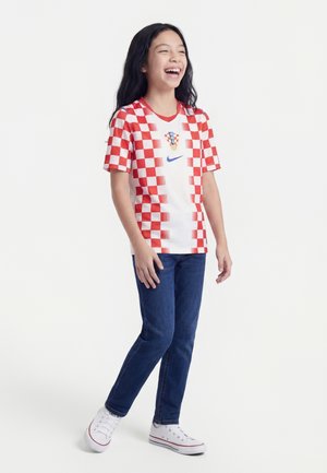 Niña sonriente, vestida con una camiseta de fútbol de Croacia a cuadros rojos y blancos, jeans azules y zapatillas blancas, de pie sobre un fondo blanco.