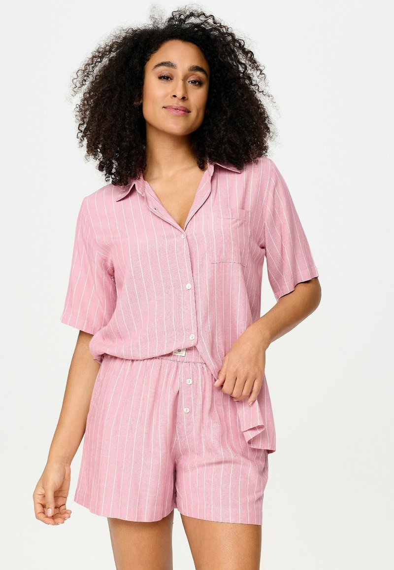 Femme aux cheveux bouclés portant une chemise à boutons rayée rose et blanche à manches courtes et un short assorti sur un fond uni.