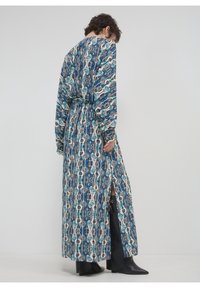 Personne aux cheveux courts portant une longue robe bleue à motifs avec une fente sur le côté, associée à des bottes noires pointues, debout contre un mur uni.
