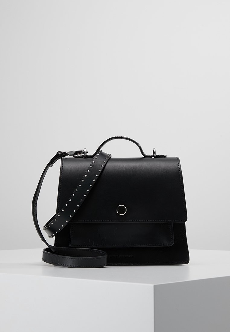 Sac à main en cuir noir avec poignée supérieure, rabat avant, détail bouton argenté et bandoulière réglable cloutée sur surface blanche.